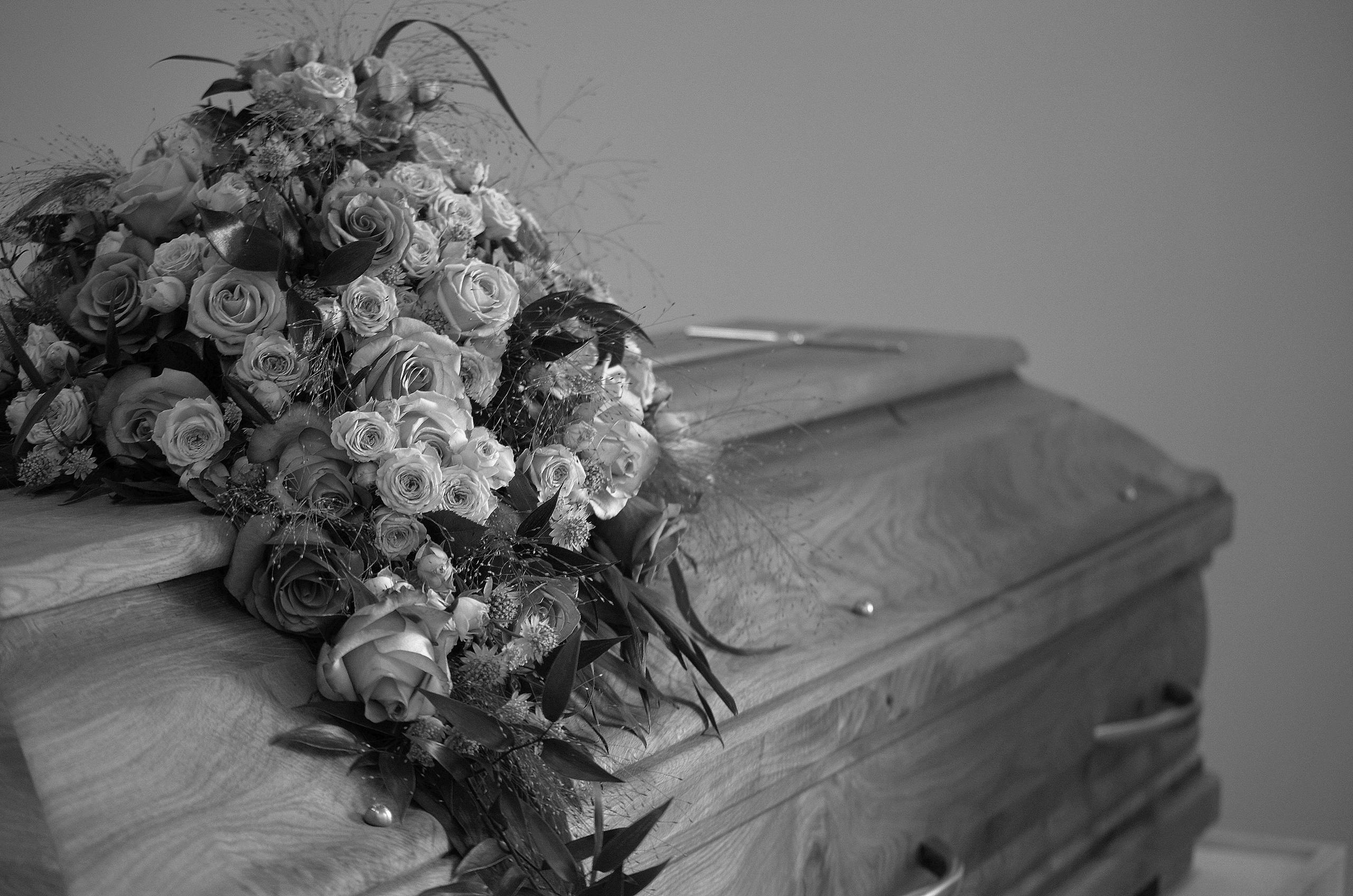 A black and white image featuring a wooden casket adorned with a vibrant floral bouquet, symbolizing respect and memory.
